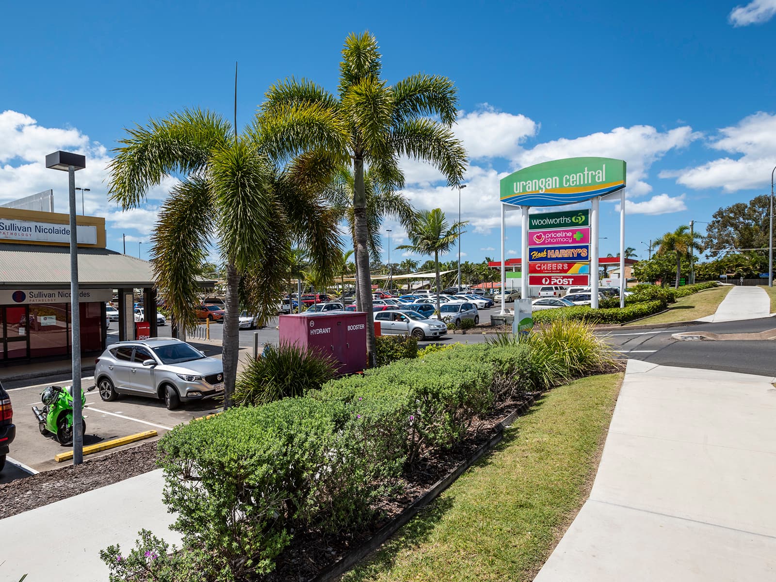Urangan Central pylon sign with Woolworths, Priceline Pharmacy, Flash Harry’s, Cheers and Australia Post, palm trees and the centre car park